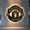 Black metal Manchester United logo wall art with bright LED backlighting on a concrete wall.