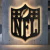 Black metal NFL shield logo wall art with bright LED backlighting on a concrete wall.