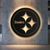 Pittsburgh Steelers black metal logo wall art with golden accents and bright LED backlighting on a concrete wall.