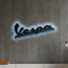 Black metal Vespa logo wall art with blue LED backlighting on a concrete wall.