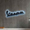 Black metal Vespa logo wall art with blue LED backlight on a concrete wall.