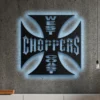 West Coast Choppers Iron Cross metal logo with blue LED backlighting on a concrete wall.